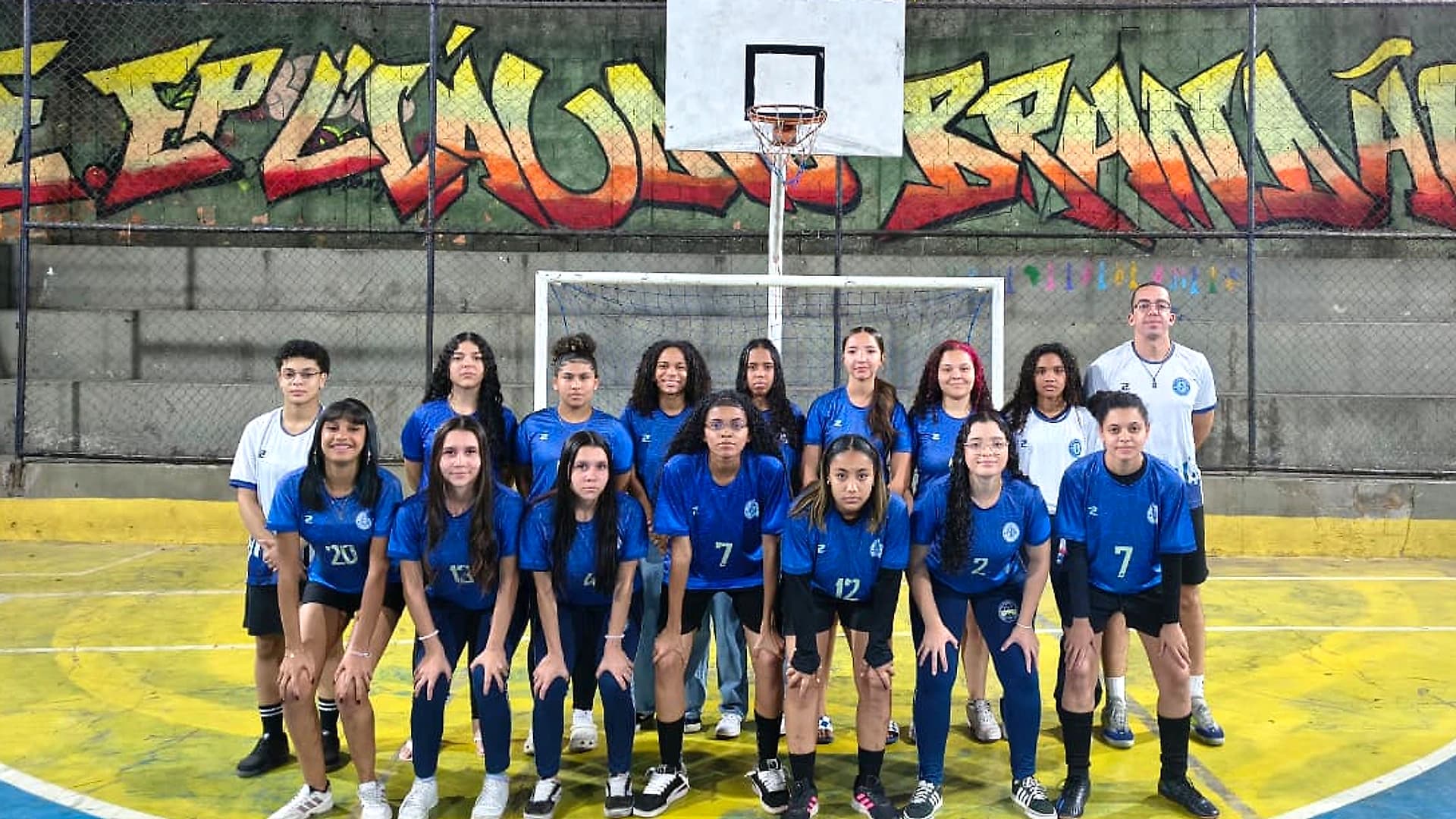 Elenco do Futsal feminino perfilado na quadra da escola, diante do gol e do grafite