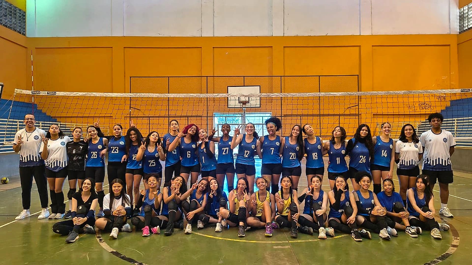 Elenco do Vôlei feminino em duas fileiras diante da rede, com a comissão técnica