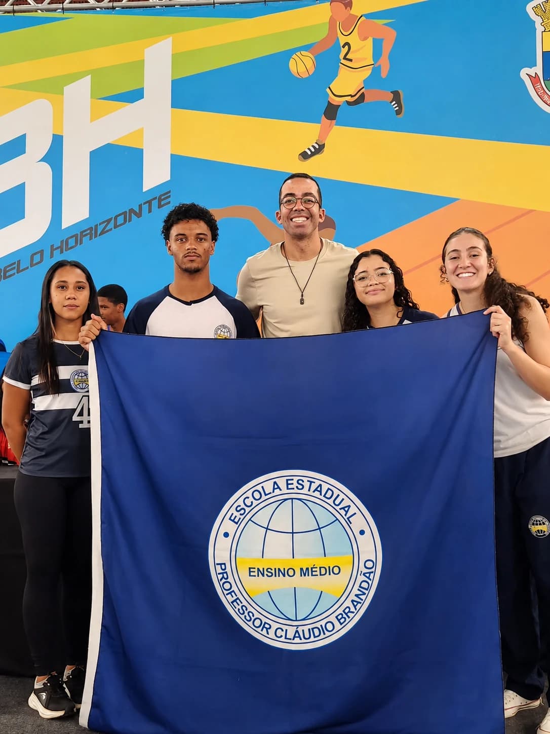 Atletas e professor com a bandeira da Escola Estadual Professor Cláudio Brandão na arena dos Jogos Escolares